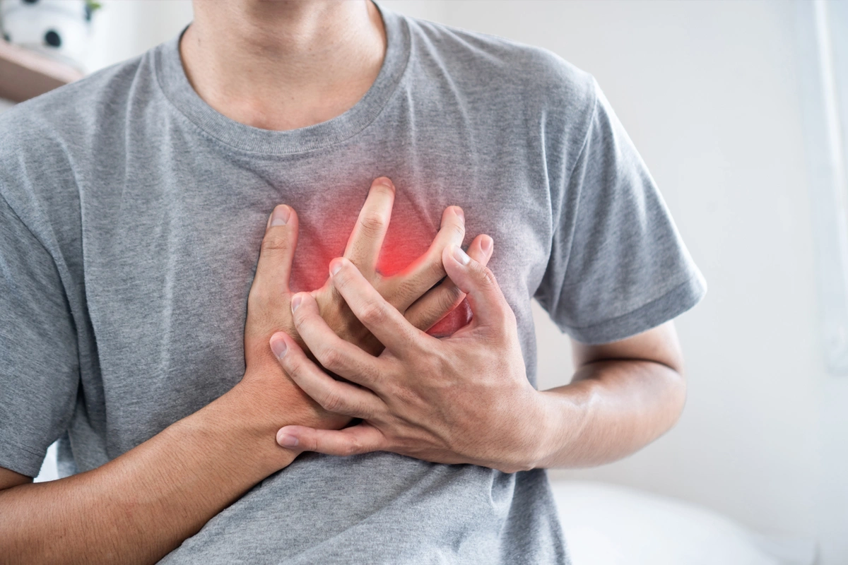 Asian man clutching his chest with both hands and wincing in pain, illustrating sudden chest discomfort or possible heart‑related symptoms.